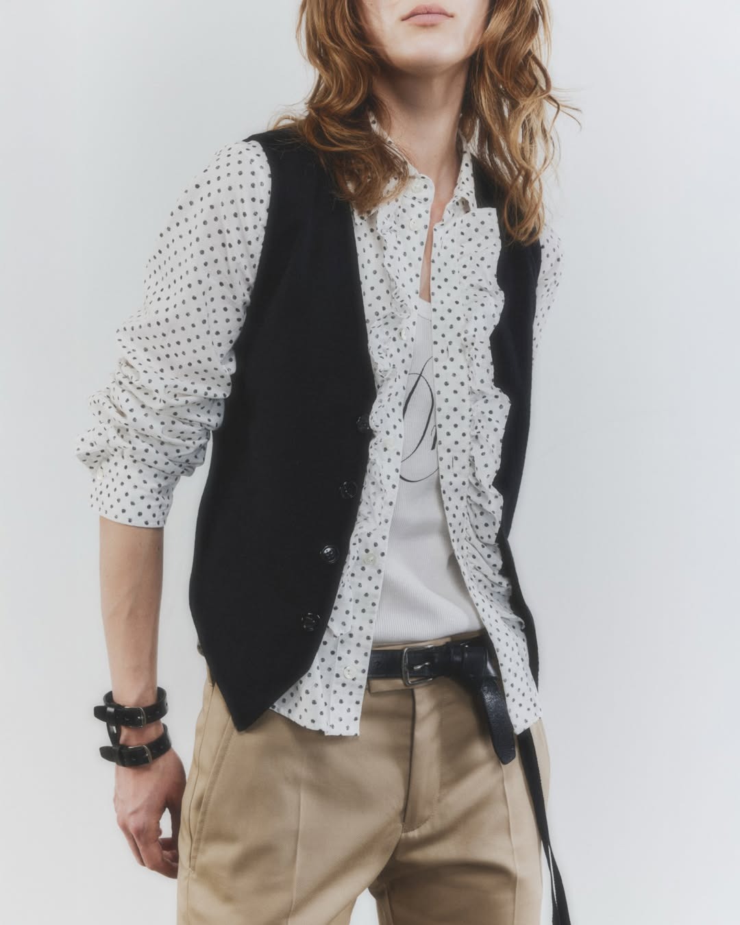 Torso portrait of person wearing white polka-dot ruffled blouse, black waistcoat, beige trousers and black leather belt