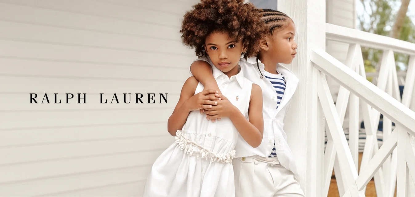 Due bambini stanno vicini. Uno indossa un vestito bianco con una cintura texturizzata; l'altro è vestito con una maglietta a righe e pantaloni bianchi. Sfondo chiaro.
