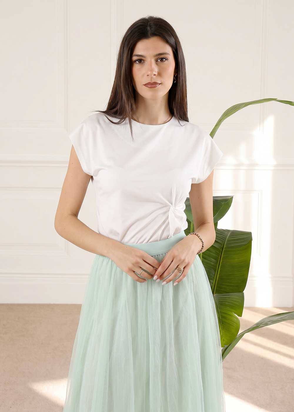 Young woman wearing white knotted T-shirt and mint tulle skirt standing indoors beside a large houseplant, hands clasped.