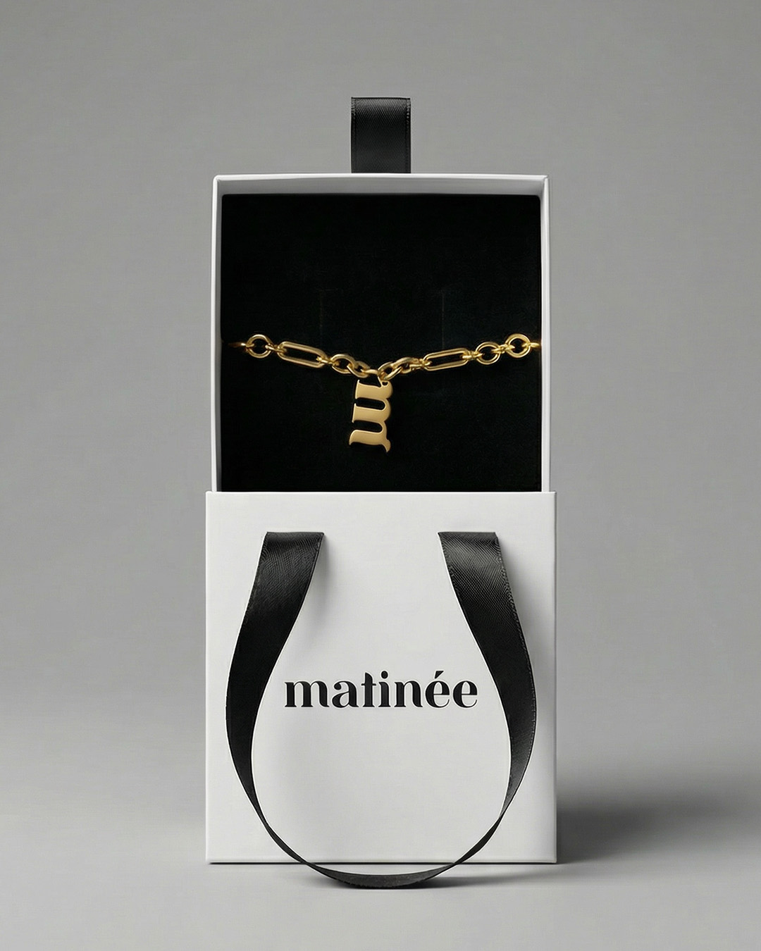 Gold chain bracelet with lowercase 'm' pendant in black-lined white gift box with black ribbon and "matinée" logo