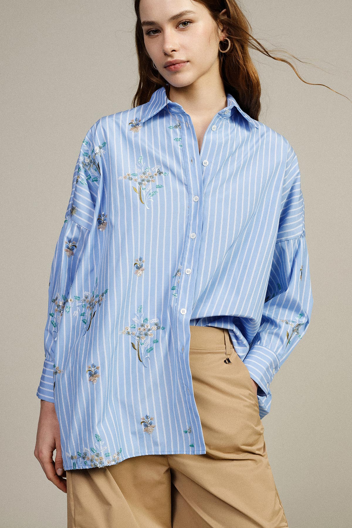 Young woman modelling blue striped embroidered oversized shirt with beige trousers against a neutral background, casual smart womenswear