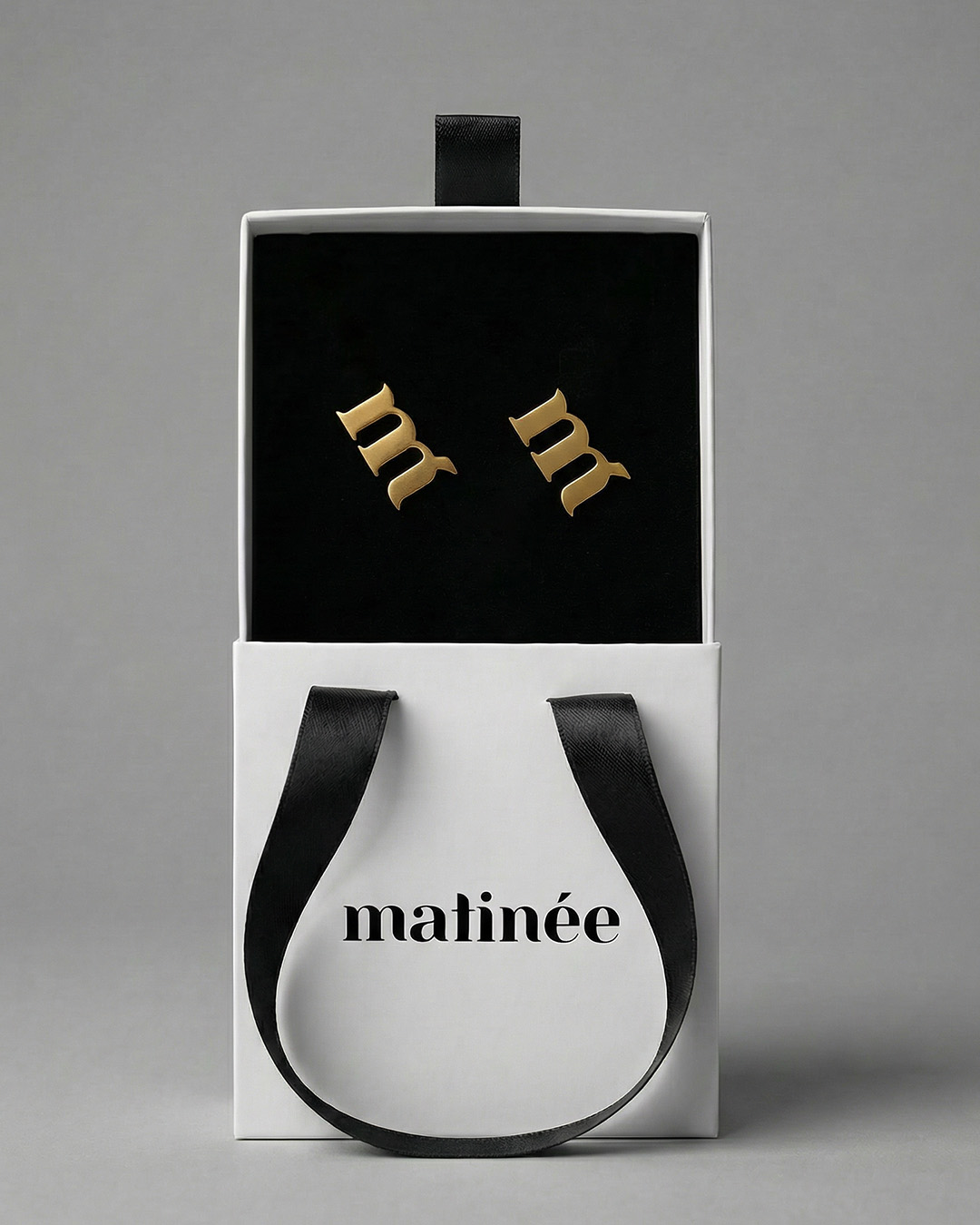 White Matinée gift box with black ribbon and velvet interior holding a pair of gold 'm' stud earrings
