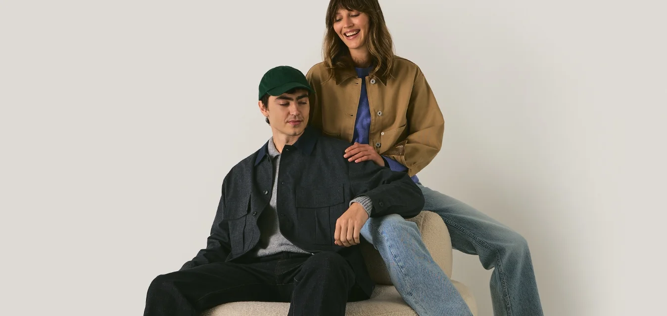 Due persone sedute su un ottomano rotondo beige contro un muro semplice; una indossa abiti scuri e un cappello, l'altra indossa jeans chiari e una giacca marrone.