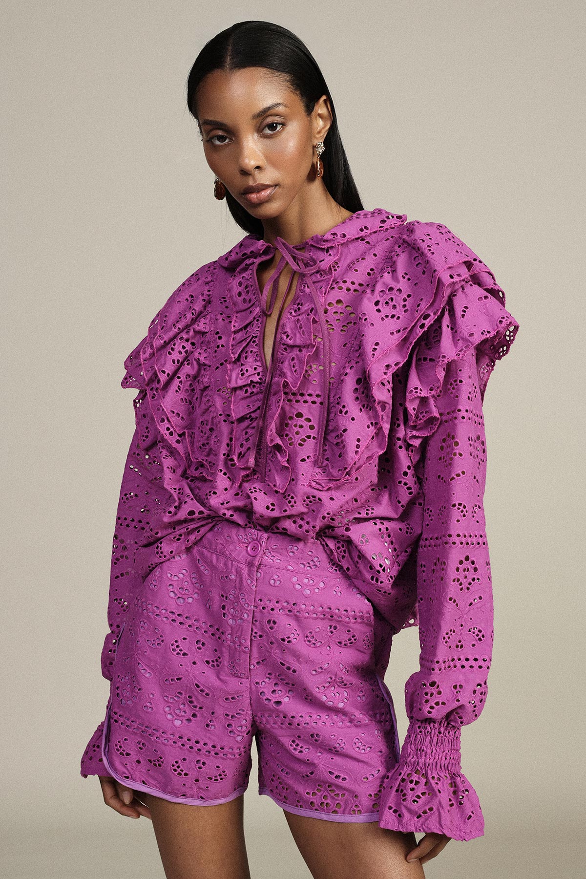 Woman modelling purple eyelet ruffle blouse with tie neck and matching high-waist shorts on neutral background