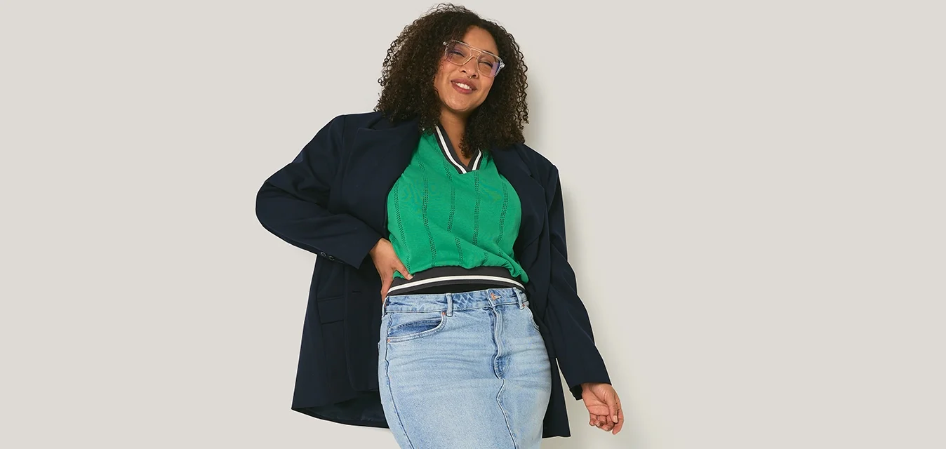 Giovane donna con capelli ricci sorridente, indossa una maglietta verde, una gonna di jeans, una giacca nera e stivali neri, in piedi davanti a un muro chiaro e semplice.