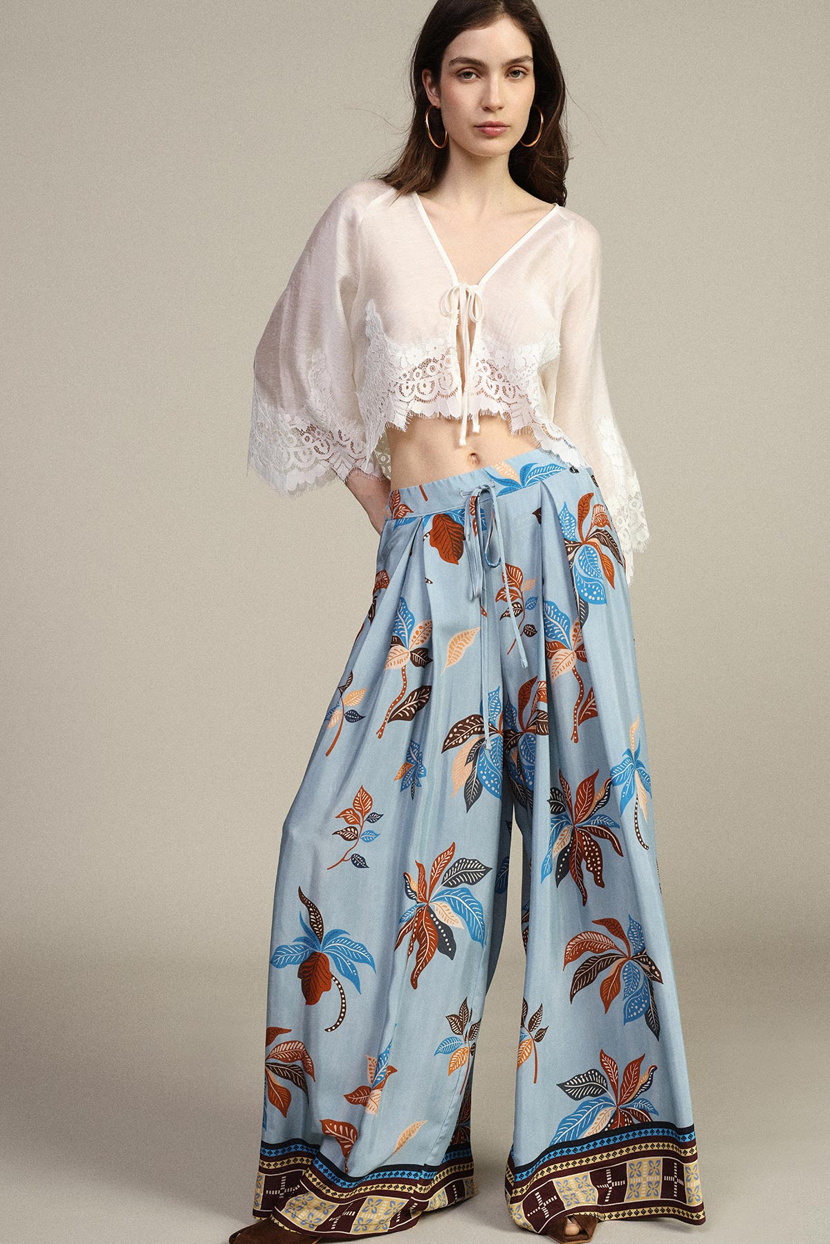 Model wearing sheer white lace tie-front cropped blouse and high-waisted wide-leg pale blue trousers with bold floral print