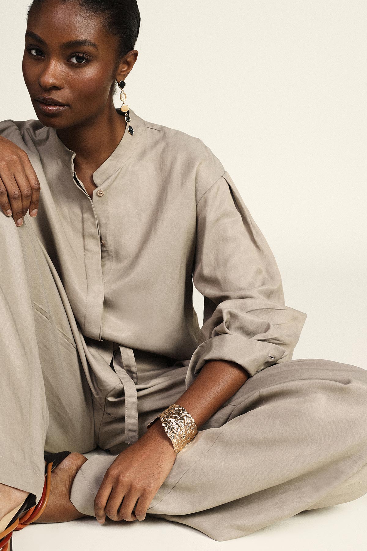 Seated model in neutral beige linen jumpsuit with rolled sleeves, gold cuff bracelet and dangling statement earrings