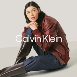 Giovane donna seduta per terra, indossa una giacca di pelle bordeaux, pantaloni blu scuro e alti stivali marrone scuro su uno sfondo chiaro e uniforme.