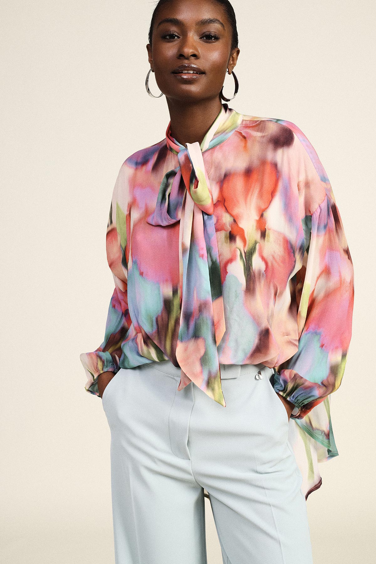 Woman wearing a colourful watercolor-print silk pussy-bow blouse paired with pale high-waist tailored trousers and hoop earrings