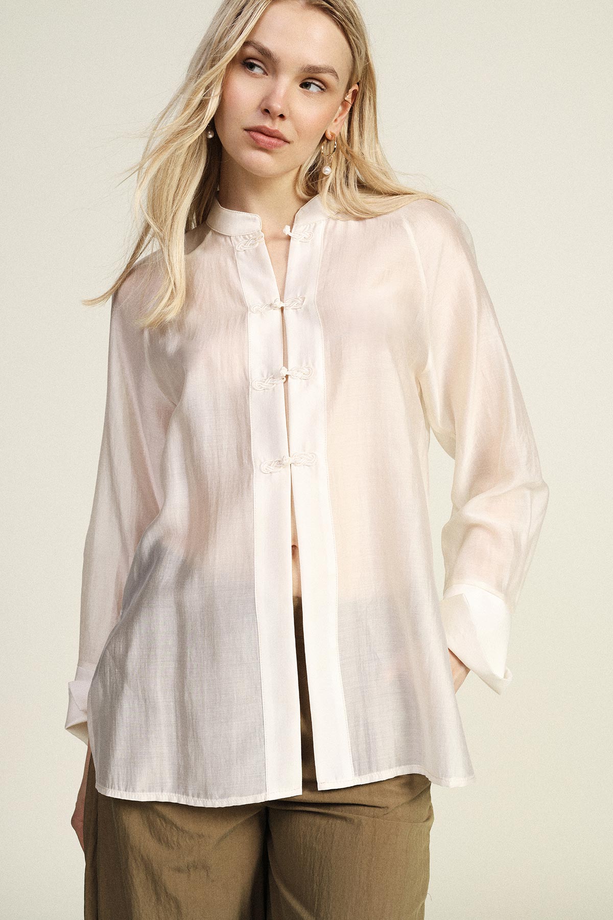 Blonde woman wearing a sheer white Mandarin-collar blouse with frog closures and khaki trousers