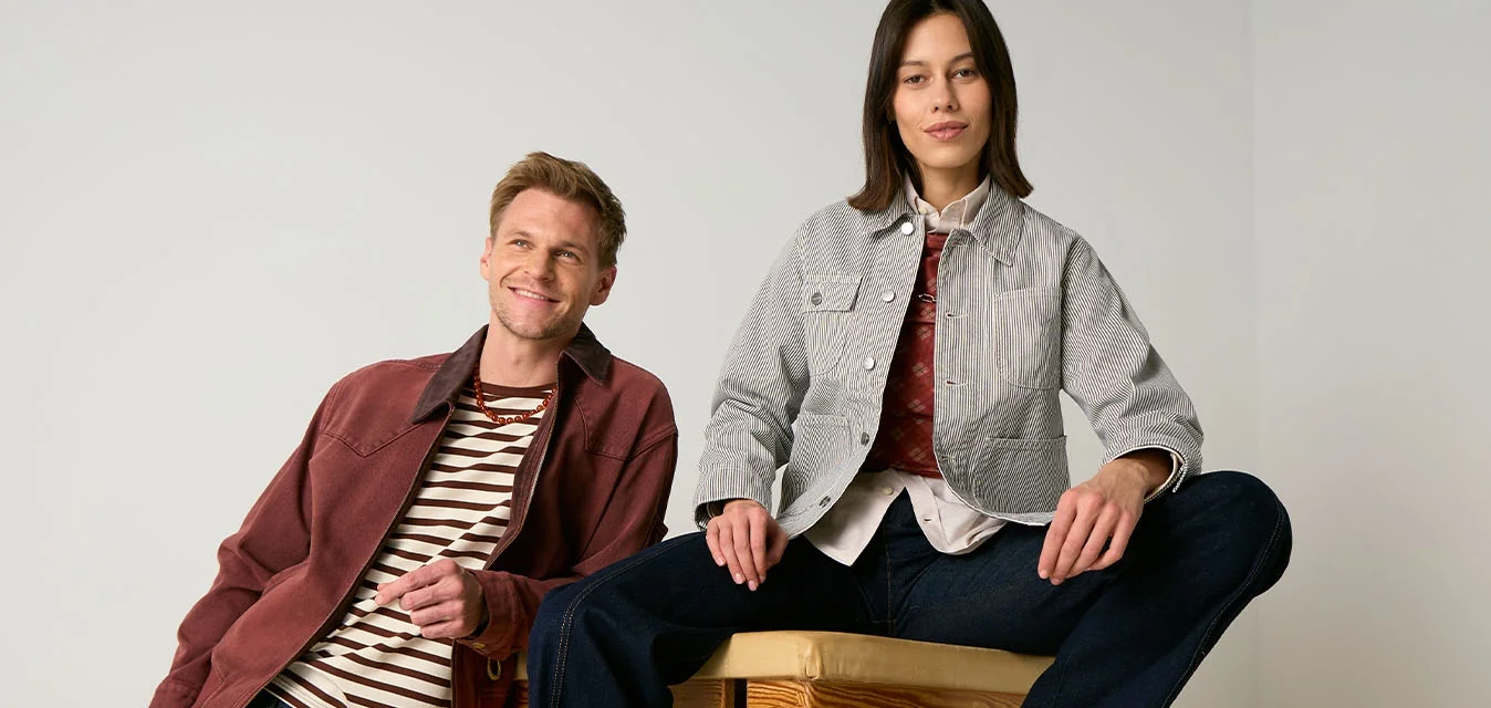 Uomo che si appoggia a una cassa di legno e donna seduta su di essa, entrambi indossano giacche casual e pantaloni scuri, su uno sfondo chiaro e uniforme.