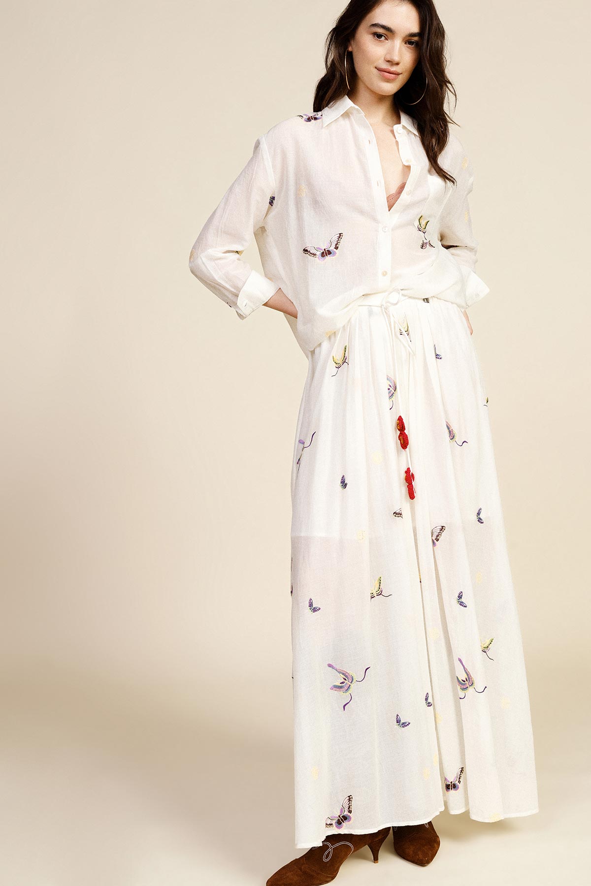 Woman wearing white embroidered button-up blouse and matching long maxi skirt with bird motifs and brown ankle boots