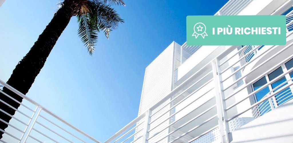 Low-angle view of a modern white seaside building with balcony railings and a tall palm tree against clear blue sky
