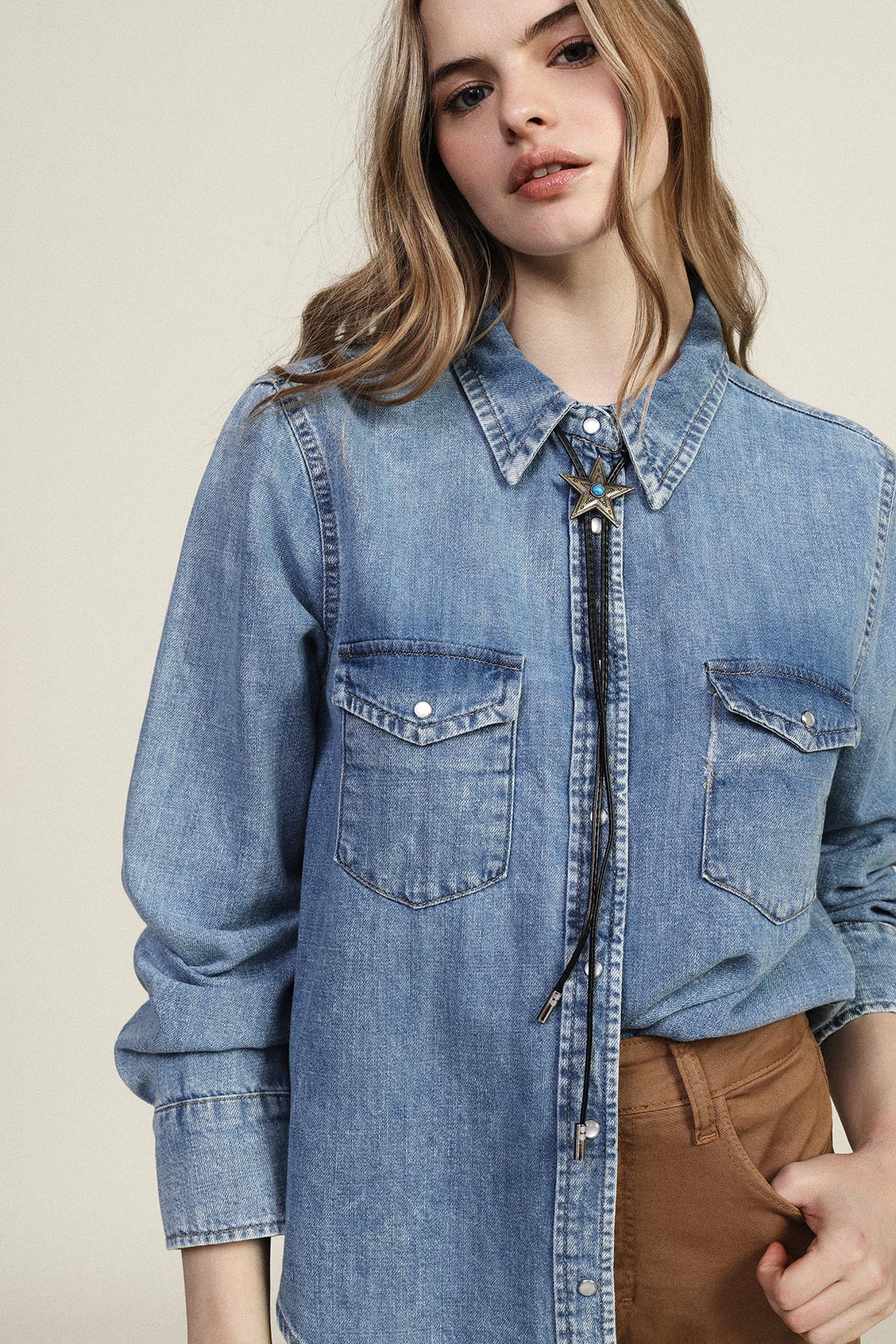 Young woman in light denim shirt with decorative bolo tie and brown trousers, hand in pocket against a neutral background