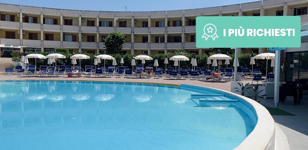 Resort swimming pool with curved hotel façade, row of sun loungers and parasols on a sunlit terrace; turquoise water and clear sky