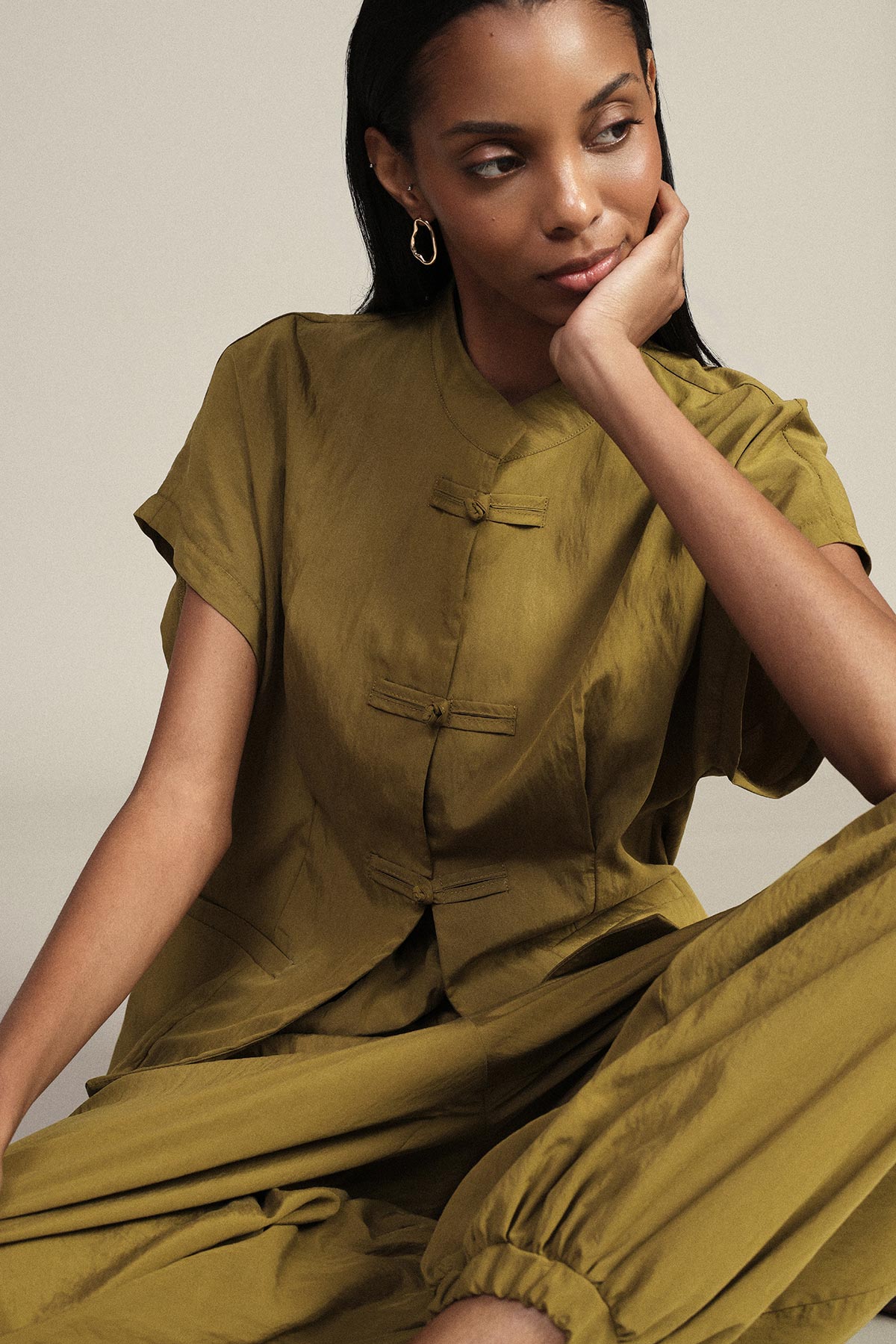 Woman seated in olive-green mandarin-collar top and matching loose trousers, resting her head on her hand, gold hoop earring