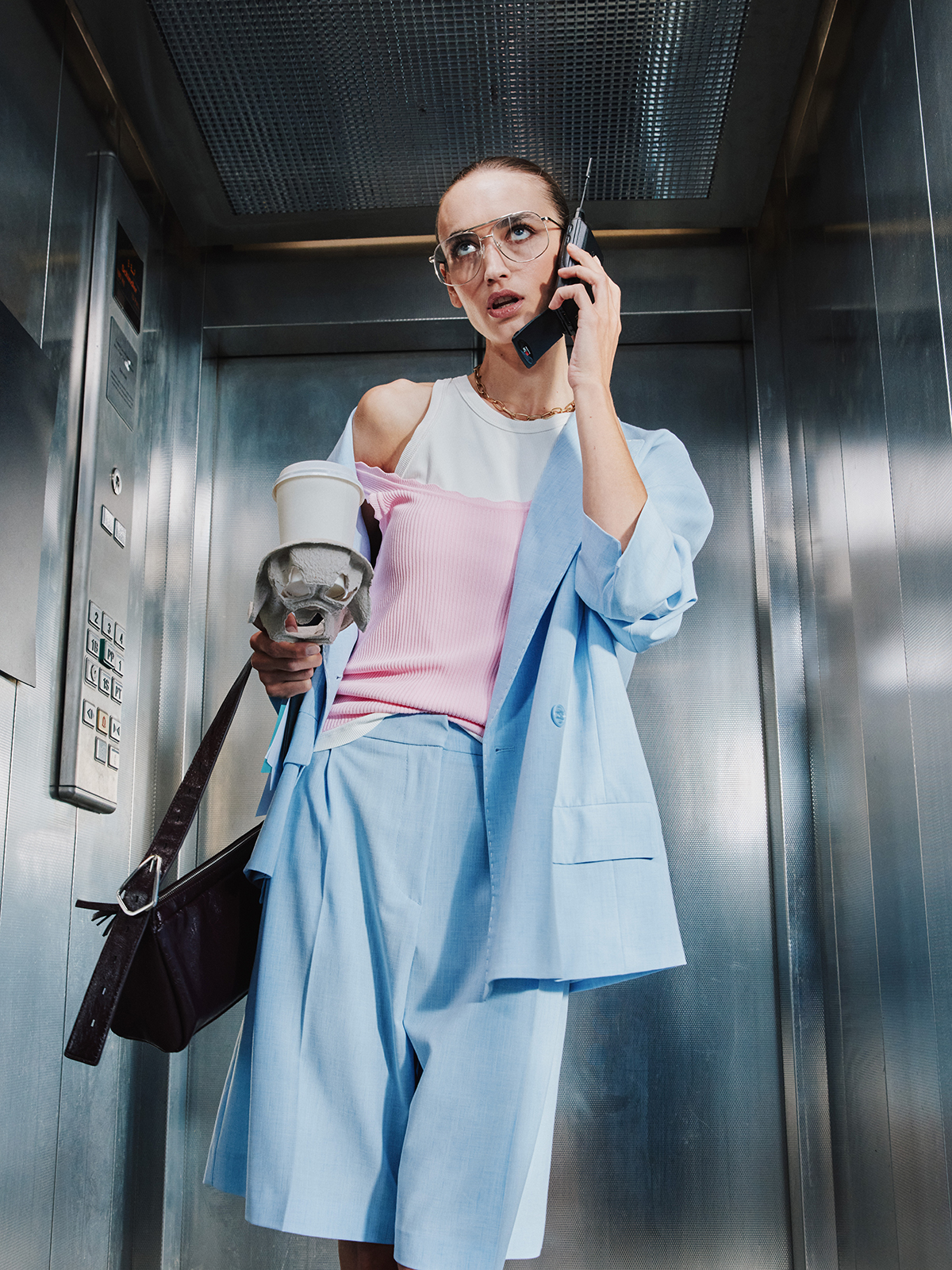 Young woman in light blue suit and pink top in a stainless-steel lift, holding takeaway coffee and talking on a phone, looking concerned