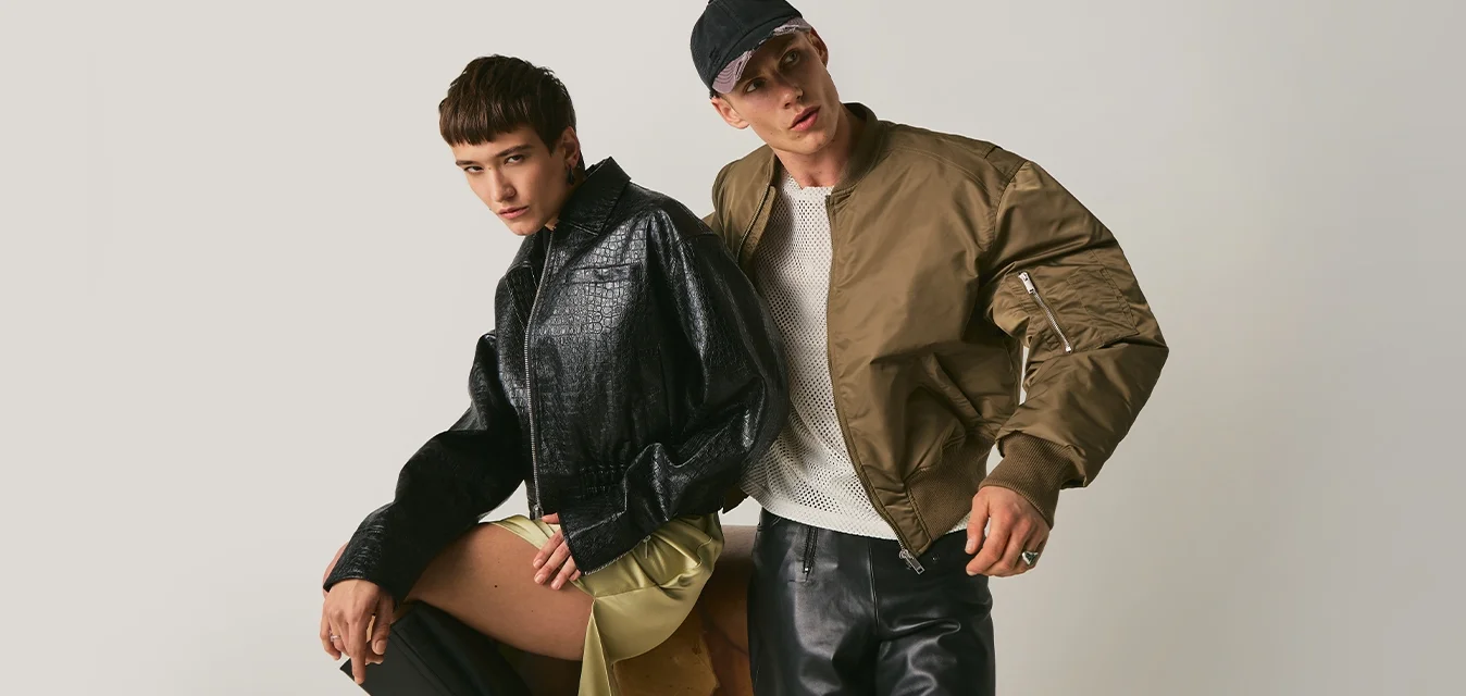 Due giovani uomini indossano giacche alla moda e pantaloni neri, posando su uno sfondo chiaro uniforme, uno seduto e l'altro in piedi con un cappellino.