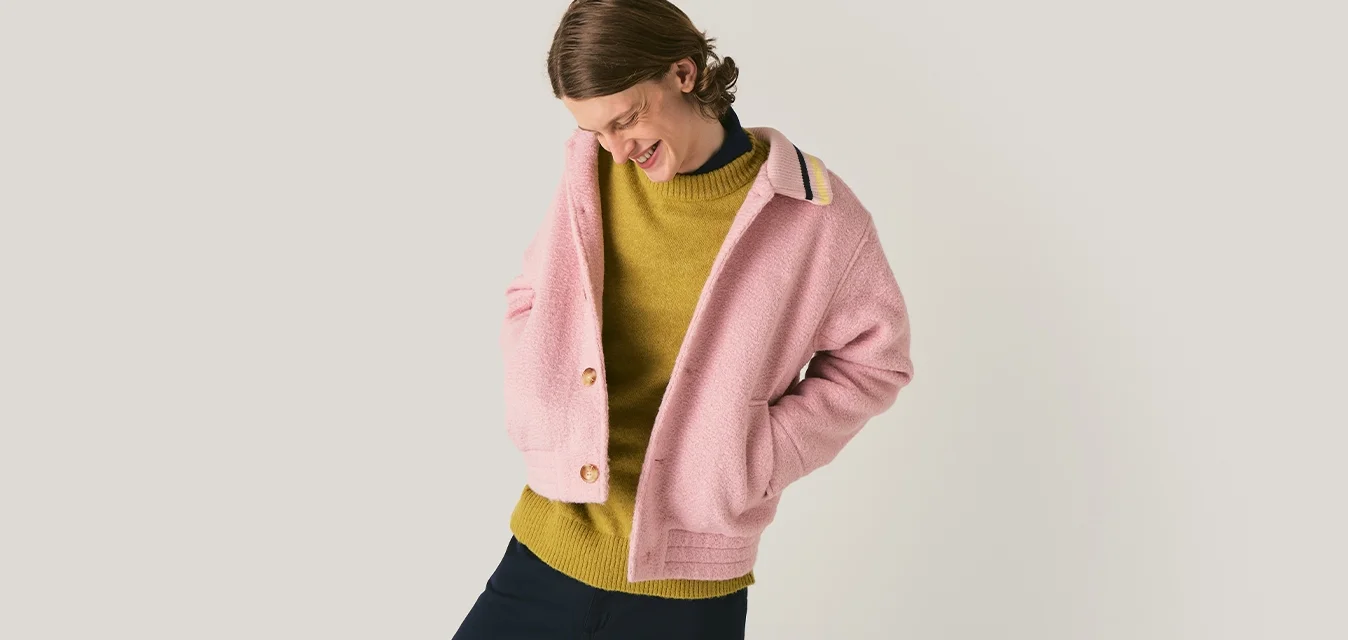 Donna che sorride e guarda in basso, vestita con una giacca rosa sopra un maglione senape e pantaloni scuri, su uno sfondo chiaro e semplice.