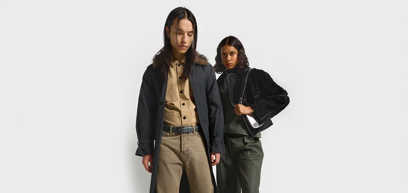 Due modelli indossano capi di abbigliamento esterno: uno indossa un lungo cappotto nero con colletto in pelliccia e chiusura a bottoni, l'altro un giubbotto corto nero textured con maniche ampie.