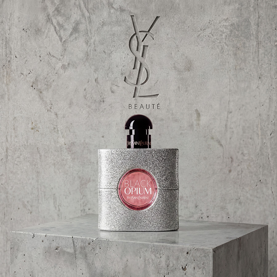 Un flacone di profumo YSL Black Opium argento scintillante su un piedistallo di pietra grigia.