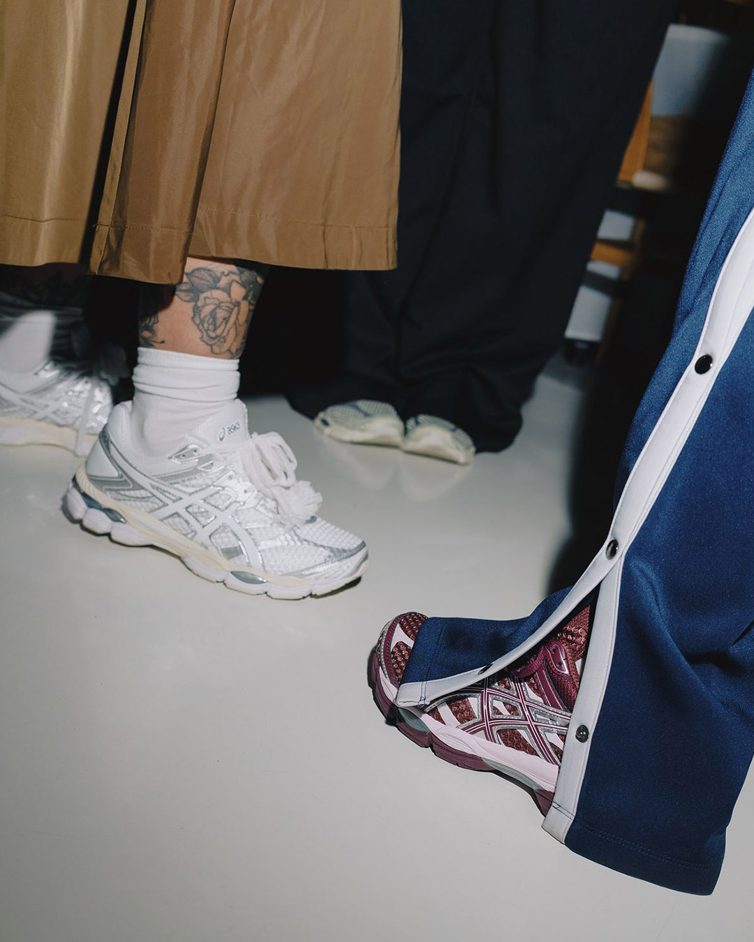 Close-up of three pairs of feet wearing Asics trainers: white mesh trainers with socks and rose ankle tattoo, maroon patterned Asics and white sliders