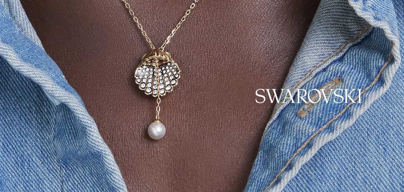 Collana con catena d'oro e ciondolo di perla a forma di conchiglia indossata su pelle scura, abbinata a una camicia di jeans azzurro chiaro.