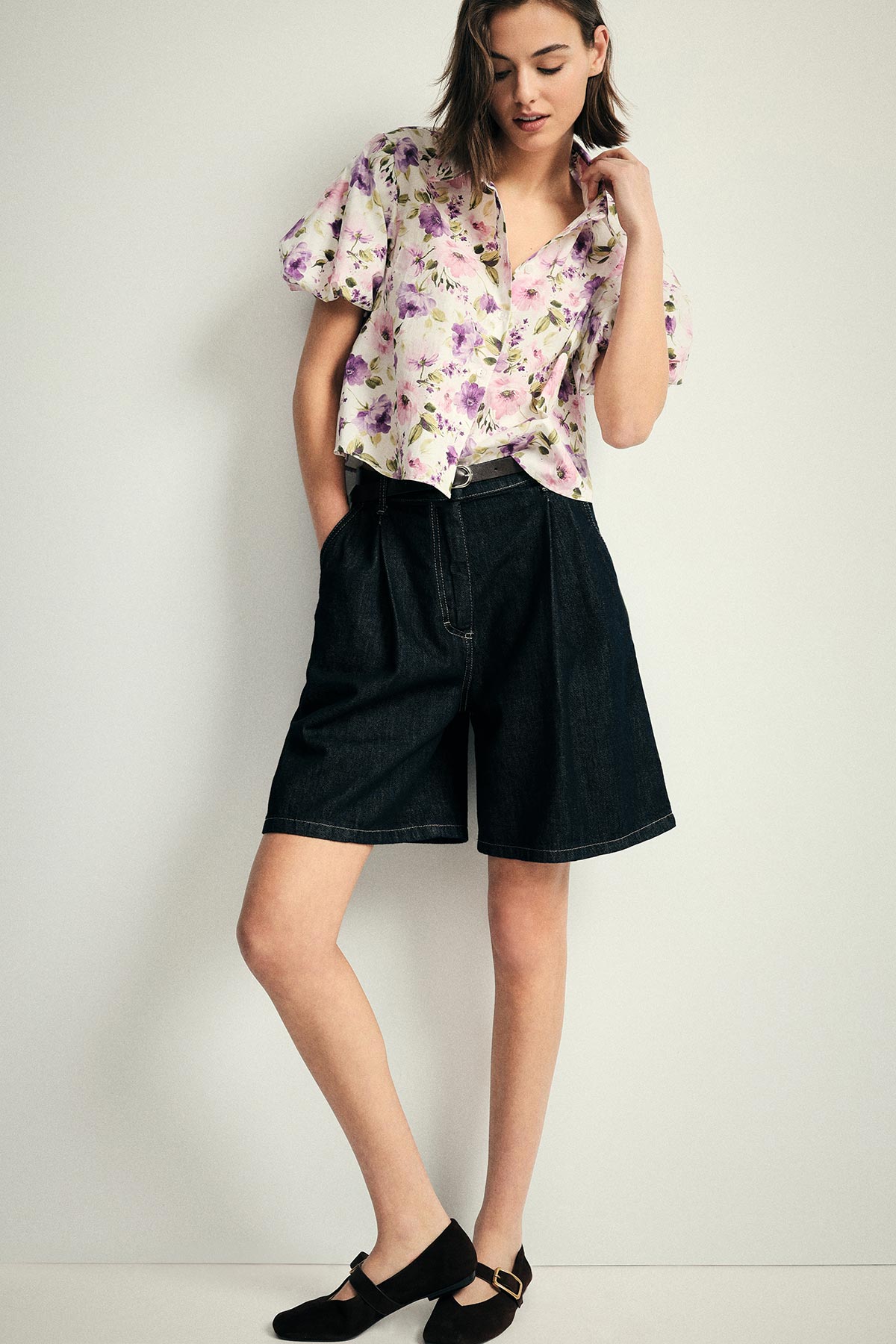 Woman in a pastel floral blouse, high-waisted dark denim Bermuda shorts and black Mary‑Jane shoes standing against a pale background