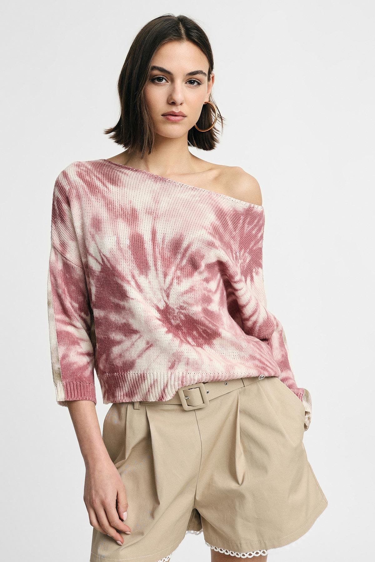 Young woman wearing an off-shoulder pink tie-dye sweater, beige belted high-waist shorts and hoop earrings against a white background