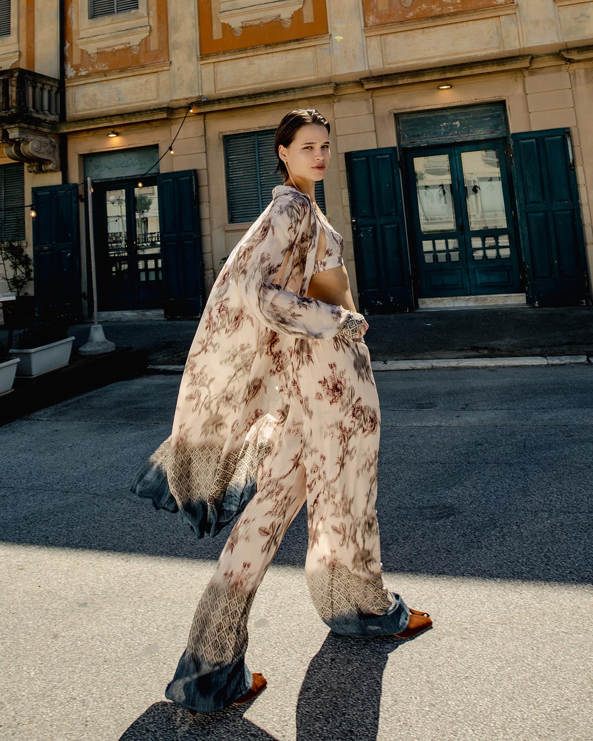 Woman walking on sunlit street in flowing floral boho outfit — matching kimono and wide‑leg trousers by vintage building