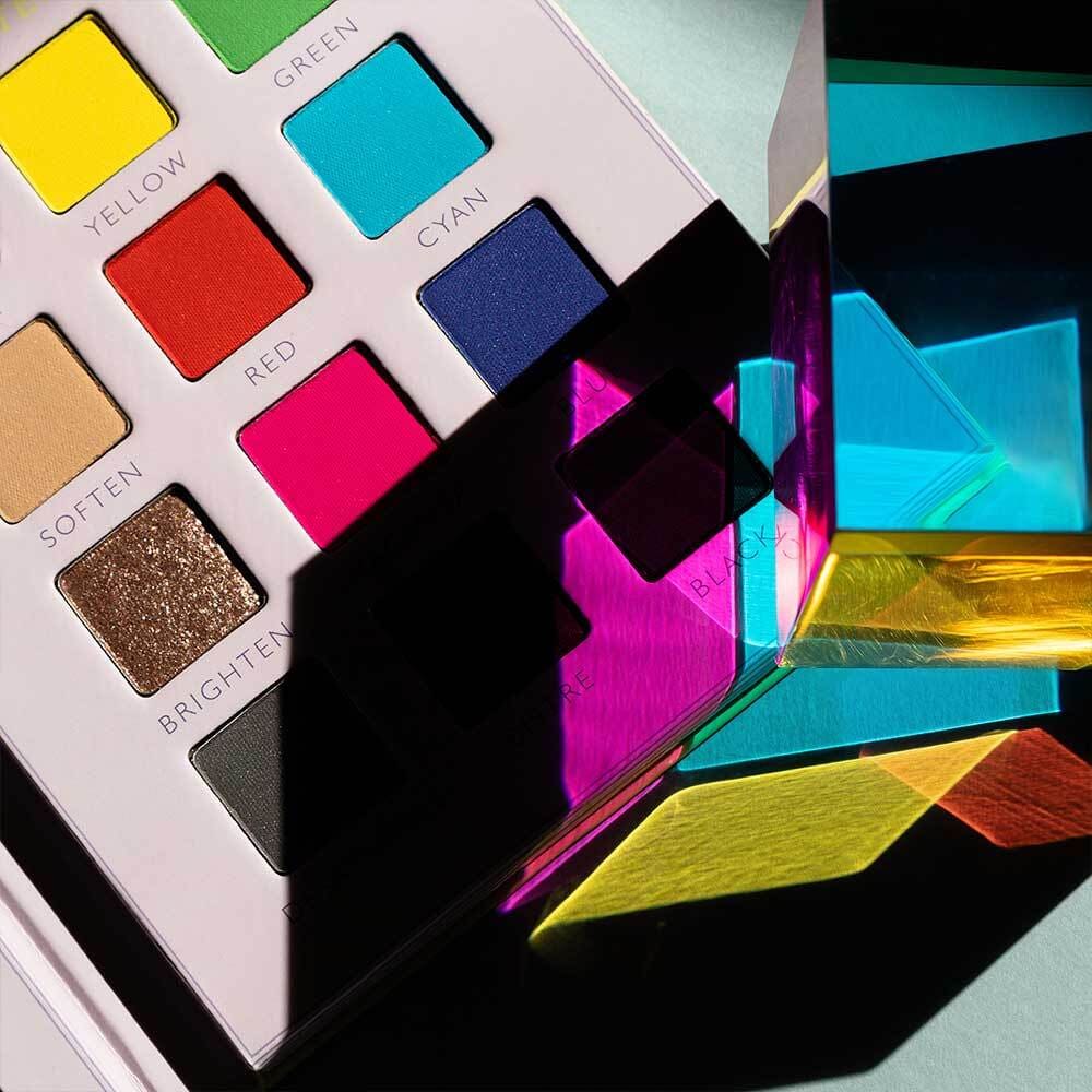 Colourful eyeshadow palette showing labelled pans (yellow, green, cyan, red, pink) with a prism casting vivid coloured reflections