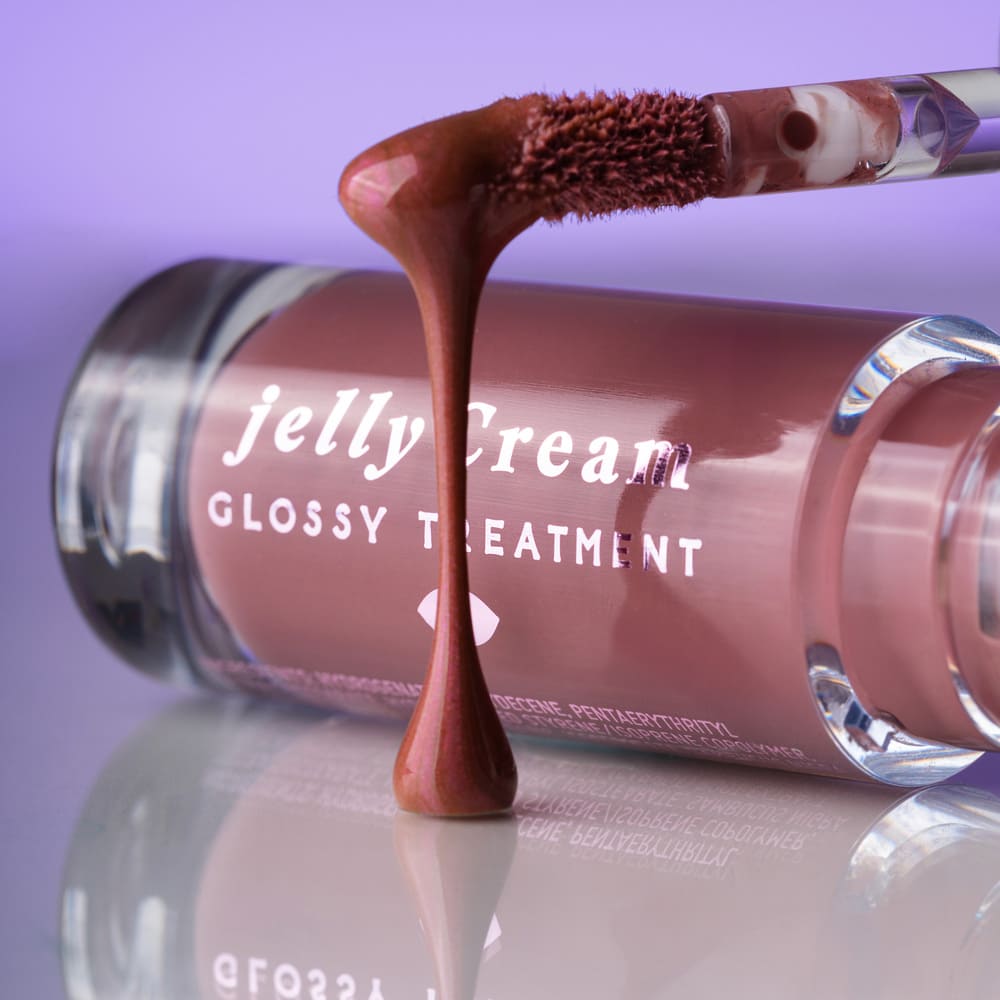Close-up of a pink-brown 'Jelly Cream Glossy Treatment' lip gloss tube with applicator dripping a glossy swatch on a reflective surface