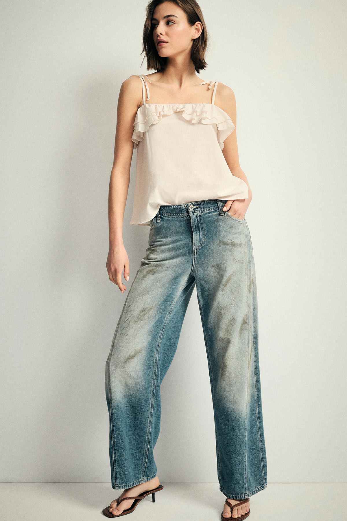 Woman modelling cream ruffle camisole and wide‑leg faded blue jeans, standing with hand in pocket and wearing sandals