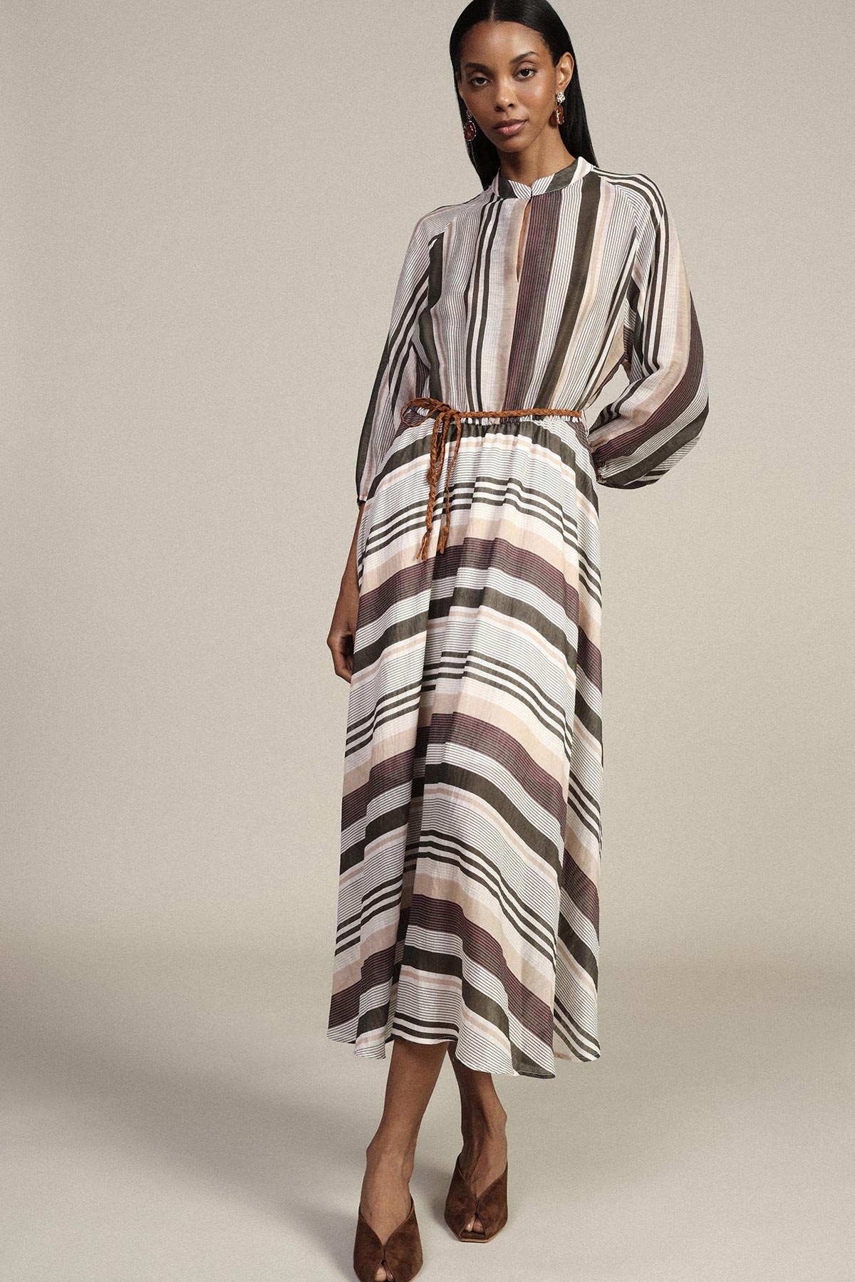 Model wearing long-sleeve striped midi dress in neutral browns with braided belt and brown heels, standing on plain studio background