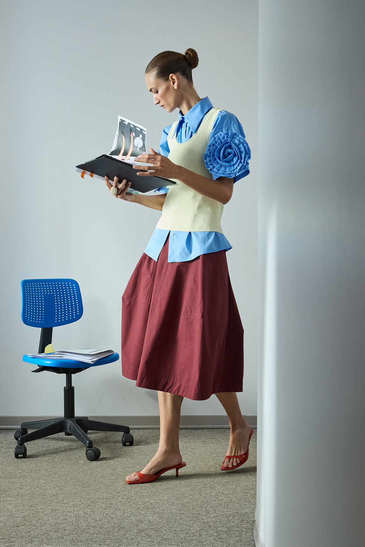 Woman in eclectic office outfit (blue blouse, yellow vest, maroon skirt) standing and reviewing documents from a black folder beside a blue chair