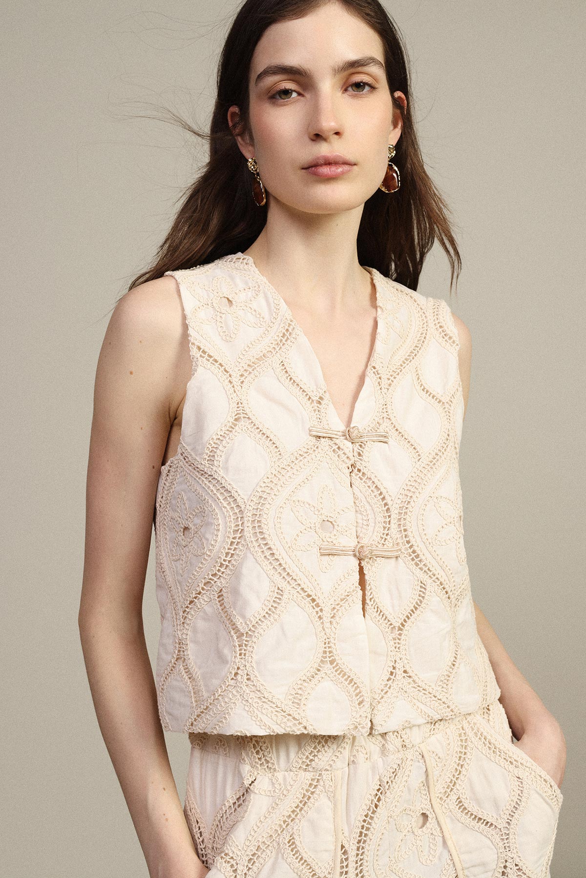 Young woman in cream embroidered sleeveless vest and matching drawstring trousers, wearing gold hoop earrings against a neutral background
