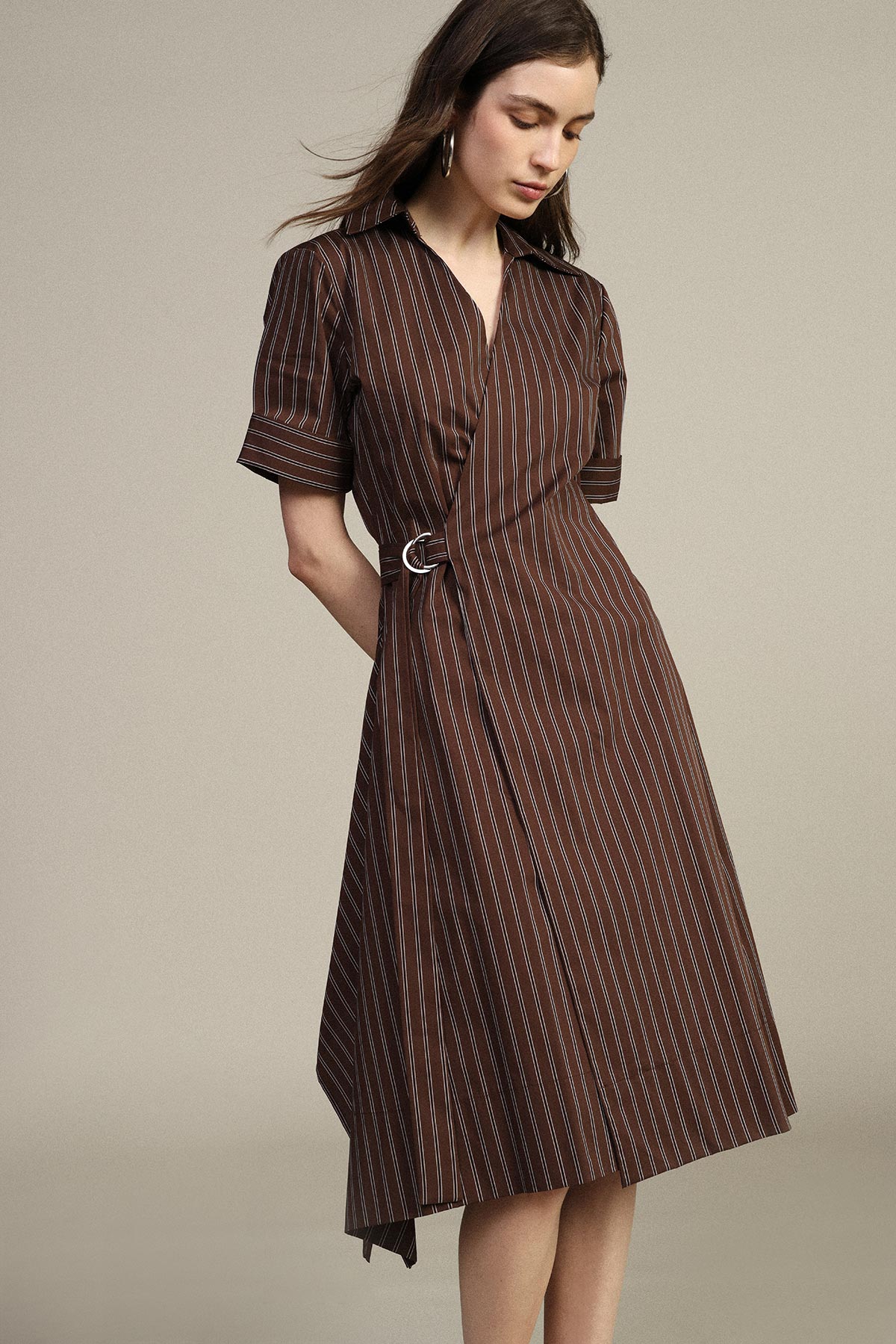 Woman in brown vertical-striped midi wrap dress with short sleeves and silver side buckle against neutral background