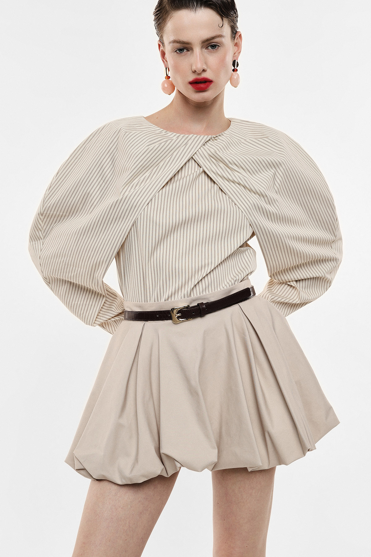 Woman model wearing beige pinstripe structured top with dramatic puffed sleeves and pleated beige mini skirt, belt and statement earrings