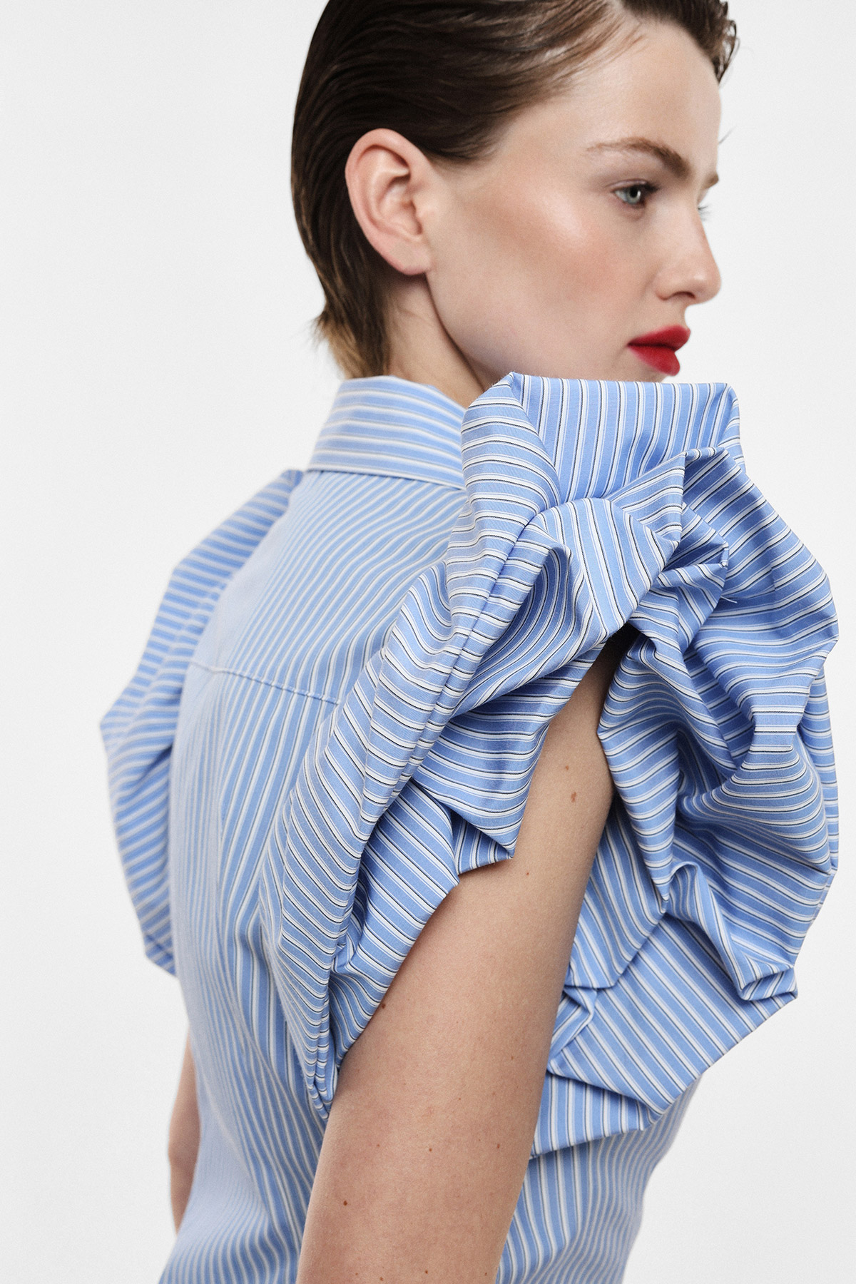 Woman in profile wearing blue striped blouse with dramatic ruffled sleeve, short slicked-back hair and red lipstick