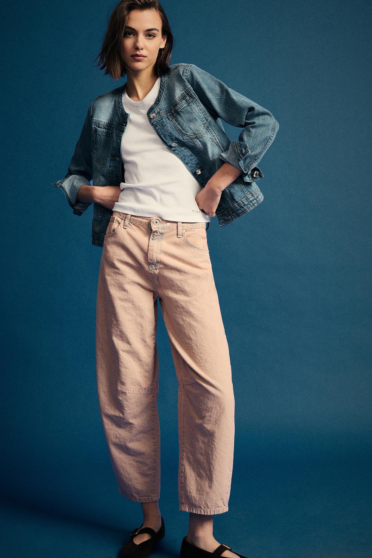 Woman wearing denim jacket, white vest top and blush wide-leg jeans with black Mary Jane shoes against blue backdrop