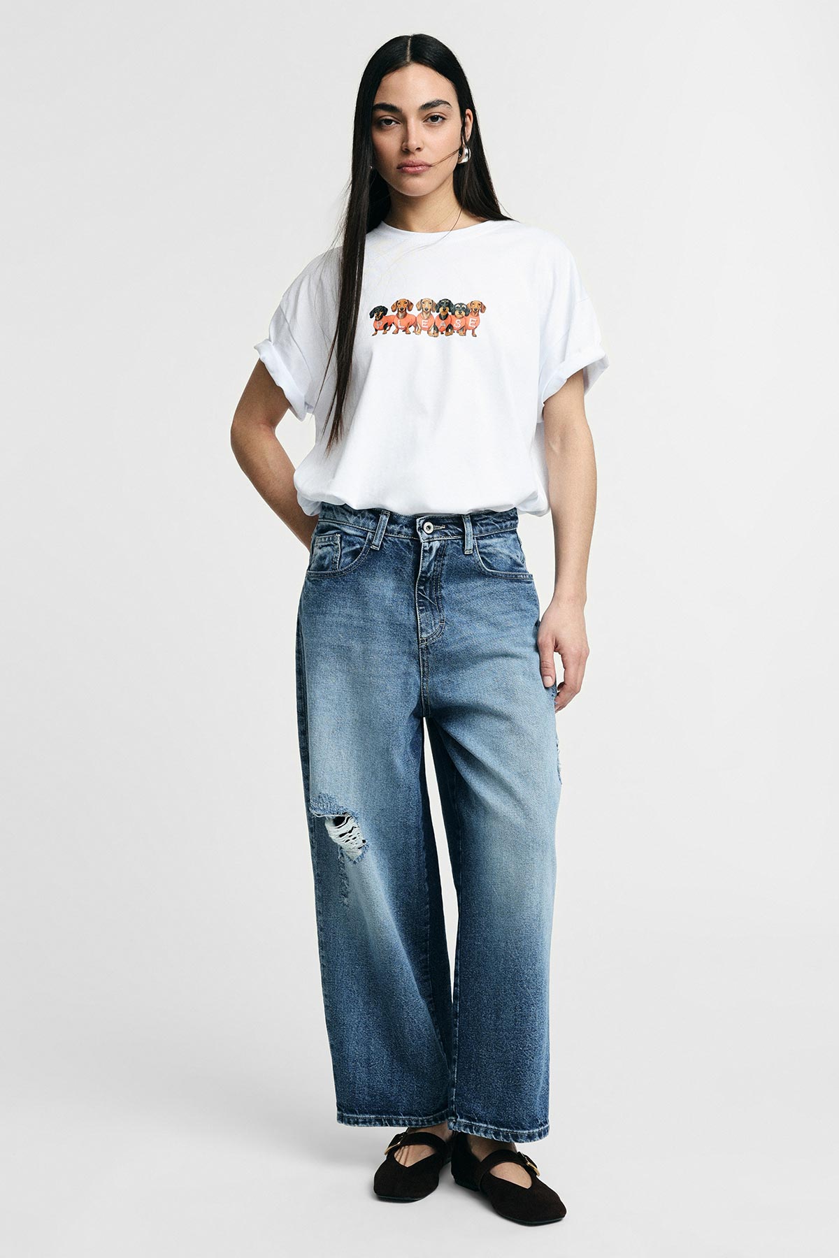 Model wearing a white graphic T‑shirt and high‑waisted wide‑leg ripped blue jeans with black ballet flats on a plain background