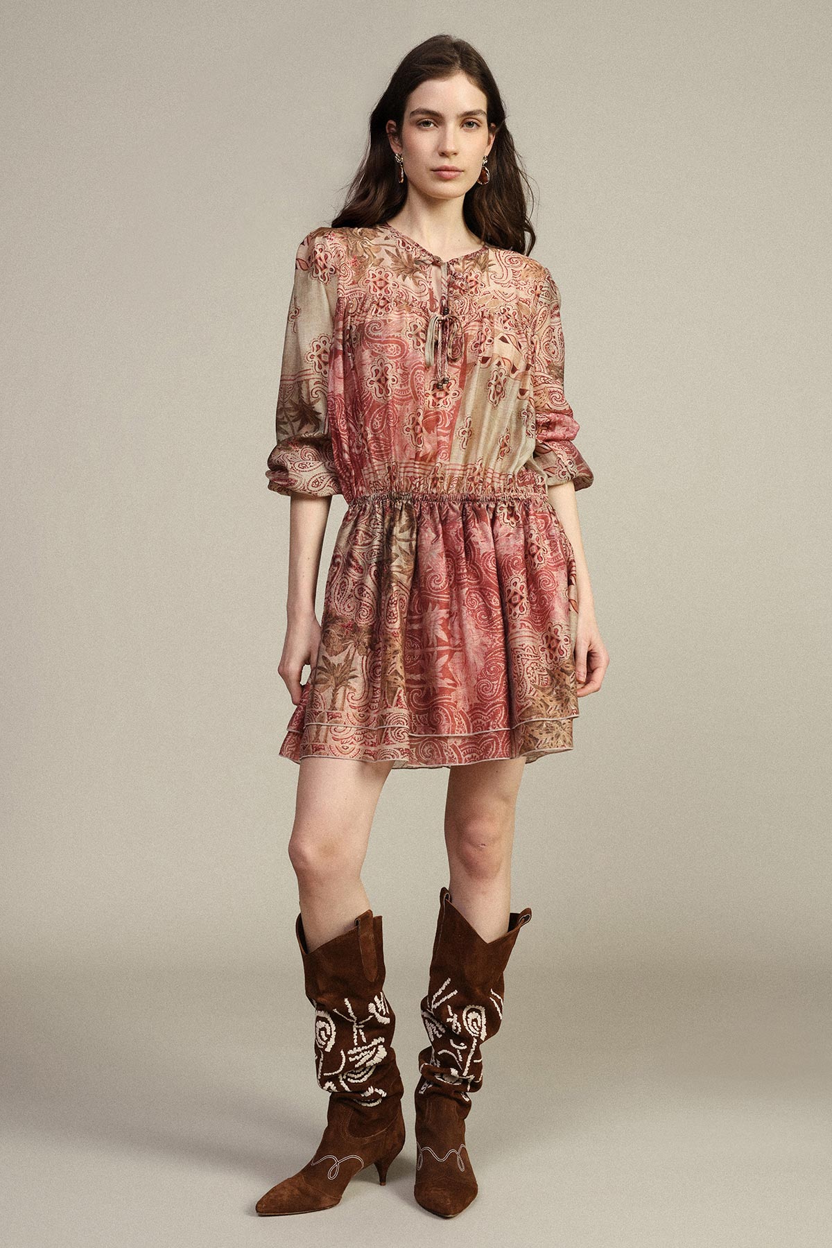 Woman in patterned pink boho mini dress with gathered waist and embroidered brown suede cowboy boots, standing