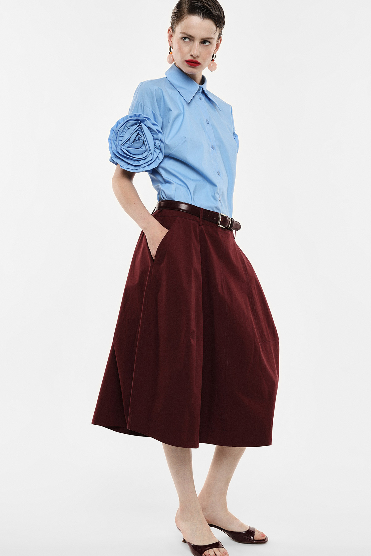 Woman wearing light-blue short-sleeve blouse with oversized rosette sleeve and burgundy midi skirt with brown belt and flats, hands in pockets