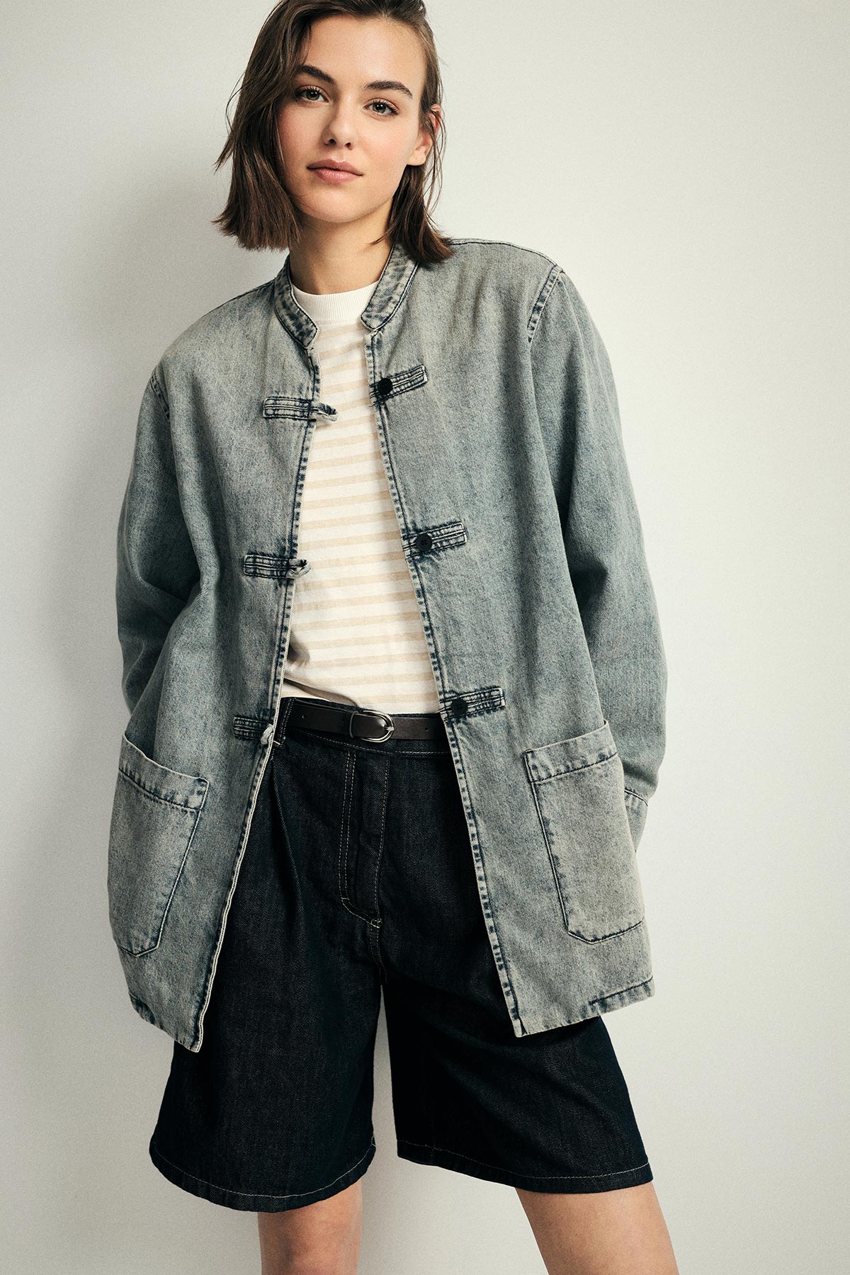 Young woman wearing oversized faded denim jacket, striped tee and black denim Bermuda shorts, standing against a pale background