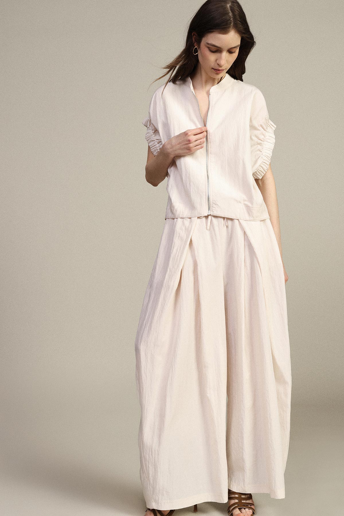 Woman wearing cream wide-leg trousers and short-sleeve zip jacket with sandals, standing against a neutral background