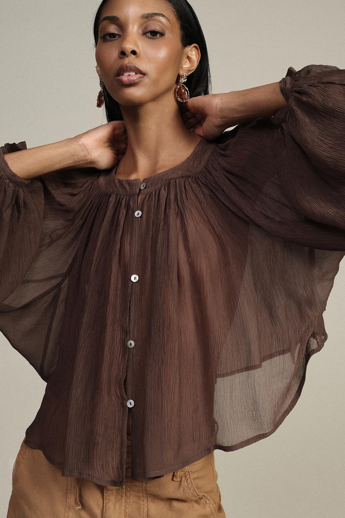 Model in sheer brown button-front blouse with voluminous sleeves and gold statement earrings, posing with hands at neck