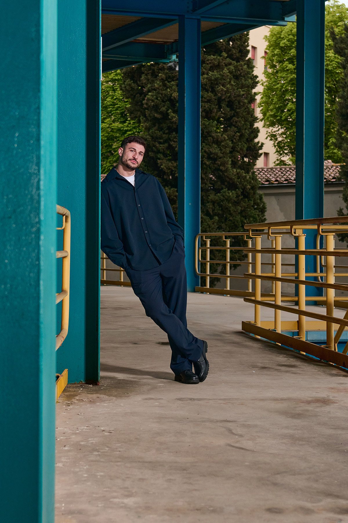 Man in dark outfit leaning against teal column on a covered walkway with yellow railings and trees in background