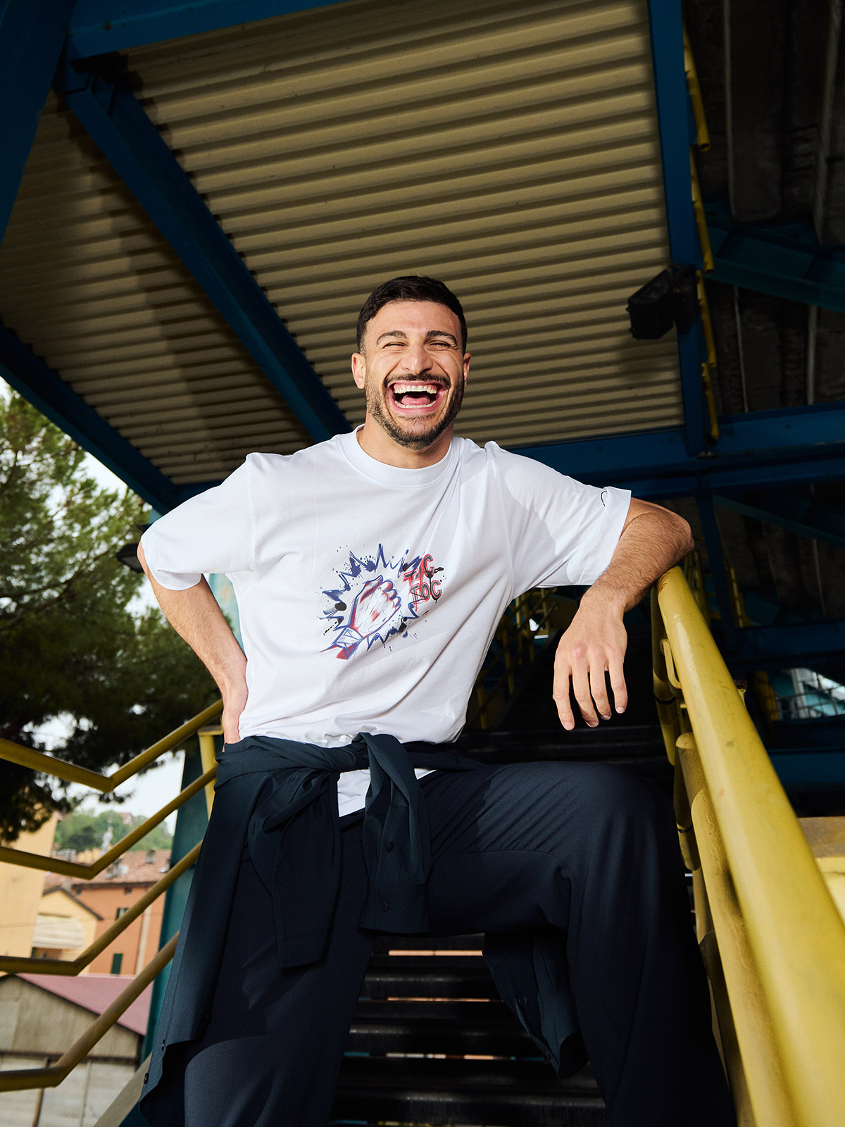 Smiling man in white graphic T-shirt and dark trousers leaning on yellow railing of outdoor metal staircase