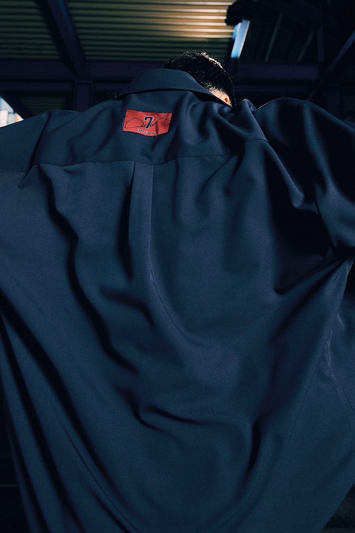 Rear view of person wearing an oversized navy coat with a red label at the collar, fabric folds and industrial ceiling in the background