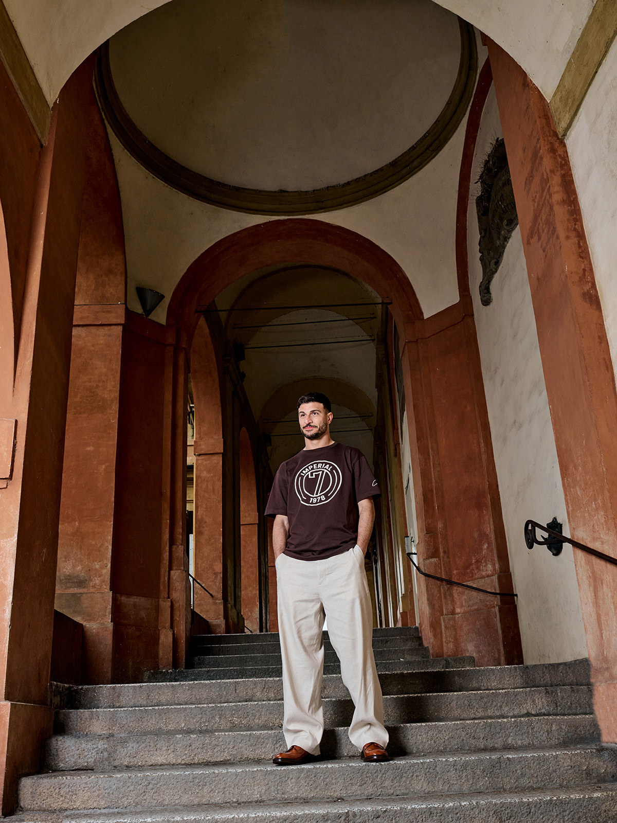 Man standing with hands in pockets on stone steps beneath a historic arched portico, wearing a brown T‑shirt and cream trousers